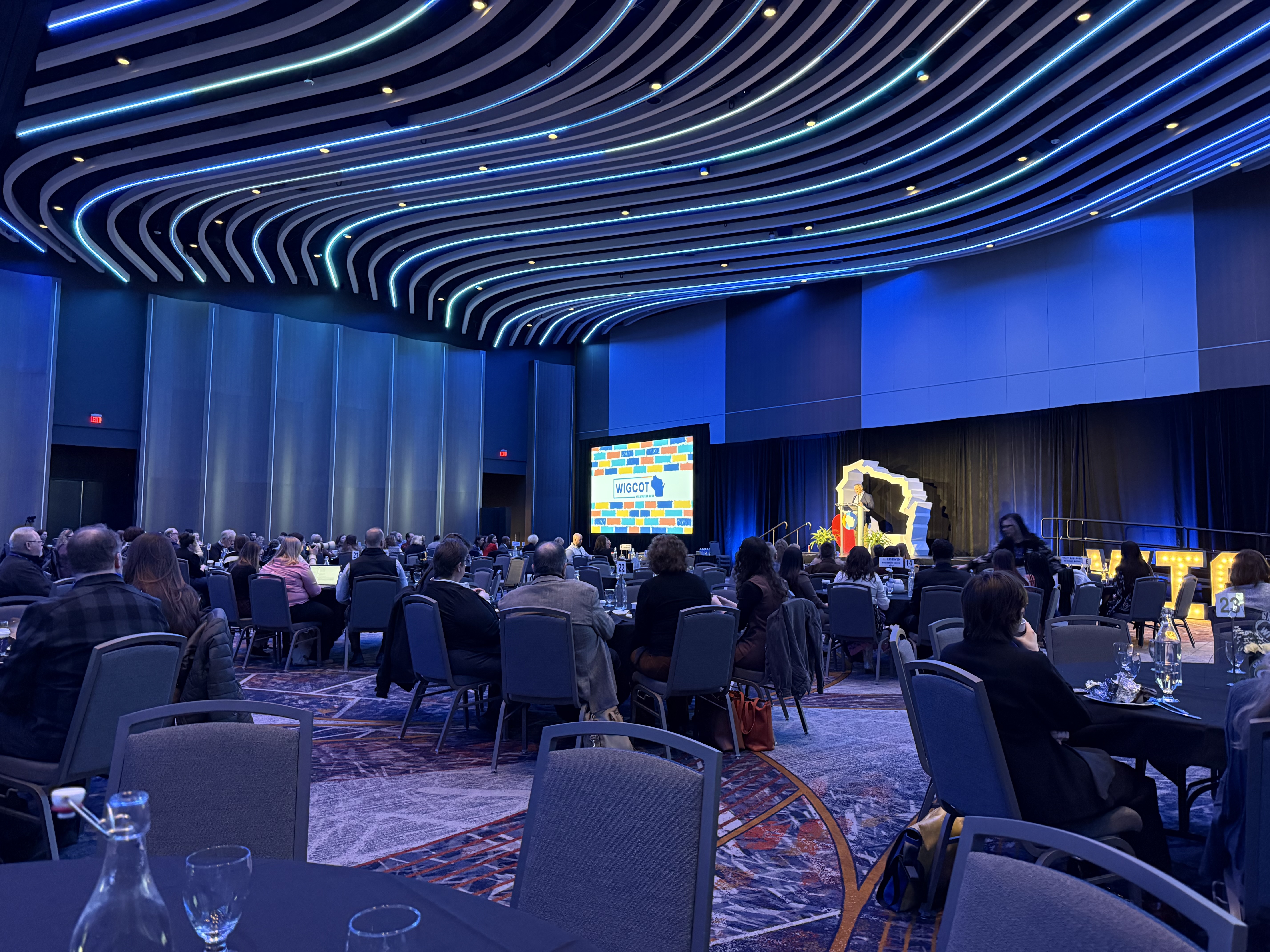 WIGCOT tourism conference — attendees at round tables, speaker at podium, blue LED ceiling architecture, authentic industry event atmosphere